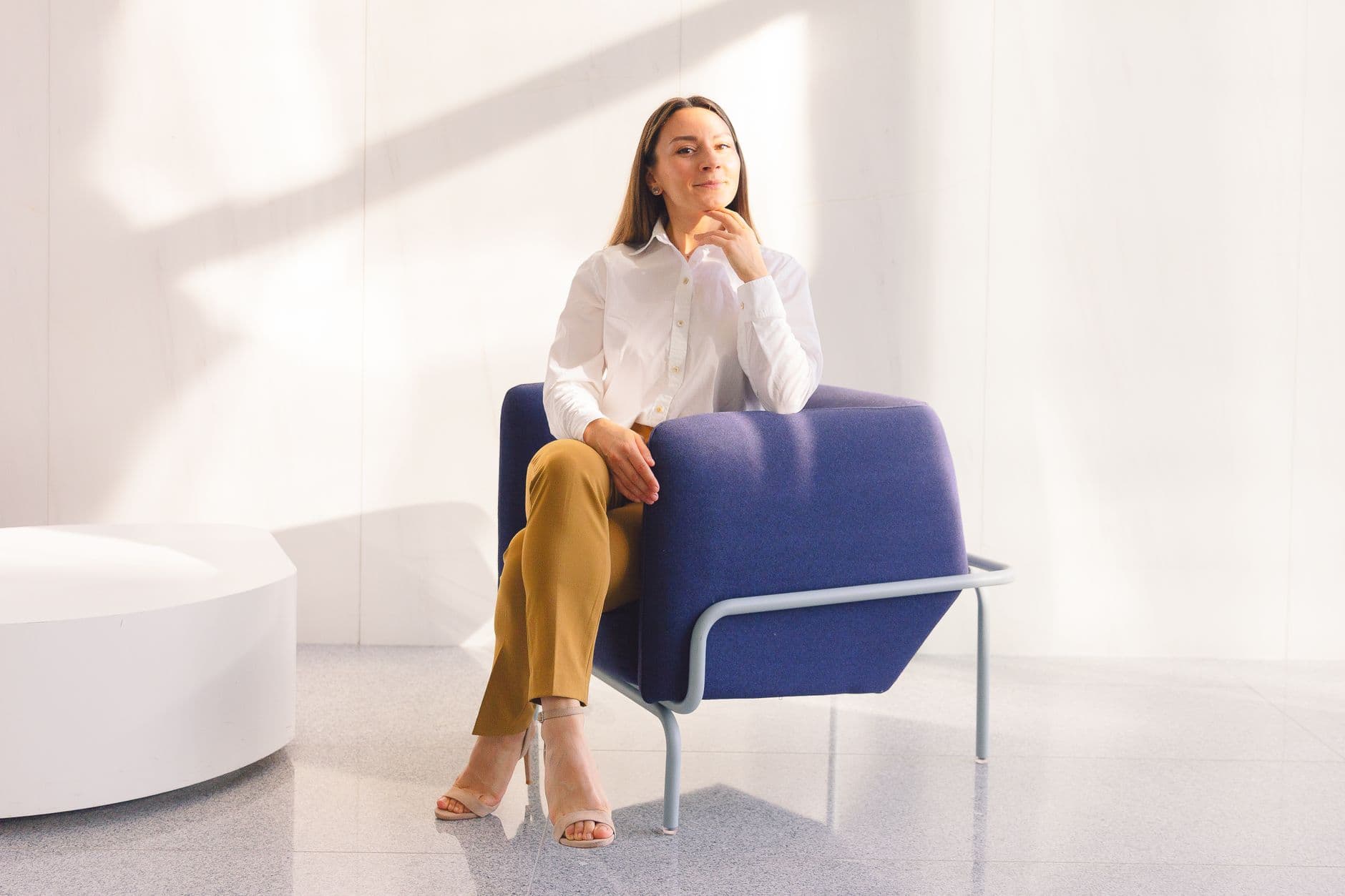 Woman in a white blouse sits thoughtfully on a blue chair in a sunlit modern office setting.