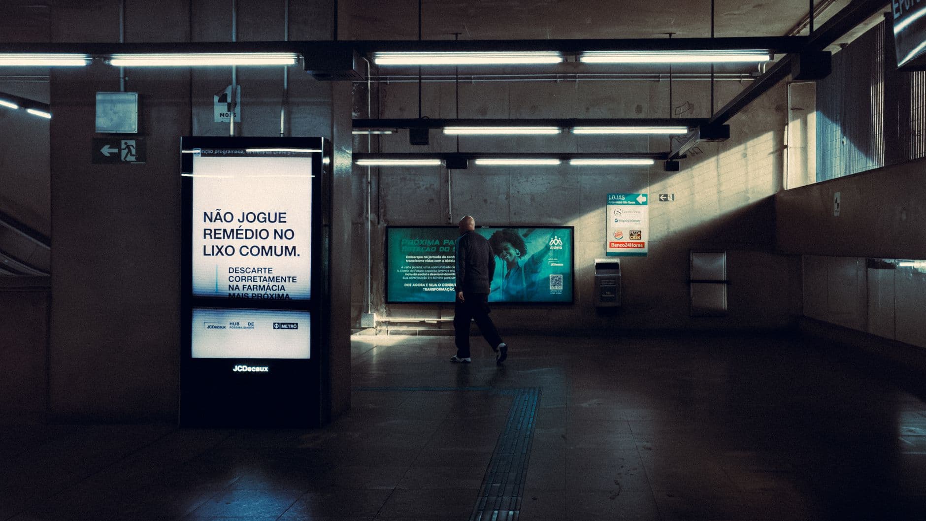 A man walks in a dimly lit modern subway station with an illuminated public service sign.